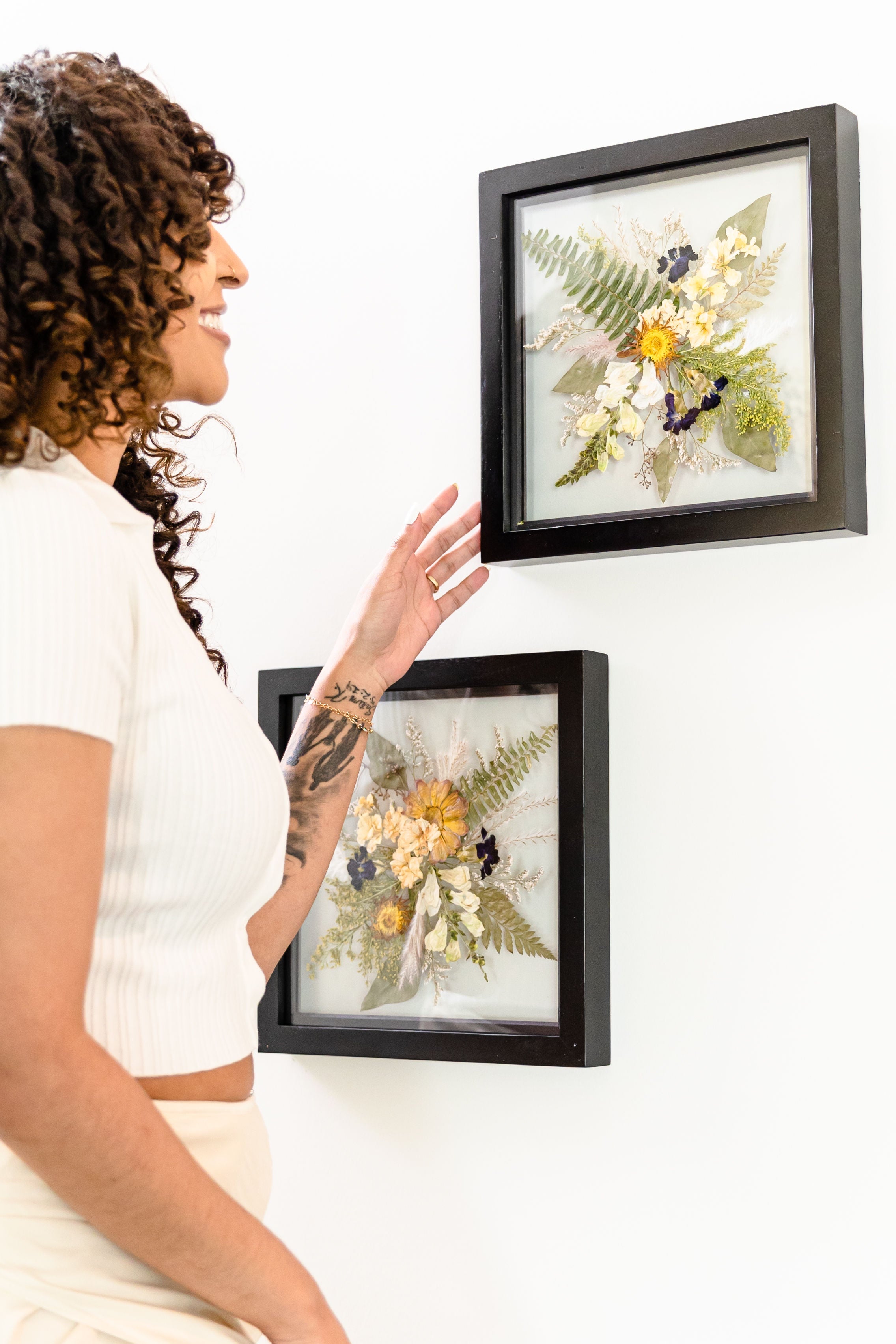 Two black float frames with pressed flowers from bouquet preservation hanging on a wall - women looks at frames as she walks by. Made by Element Design Co.