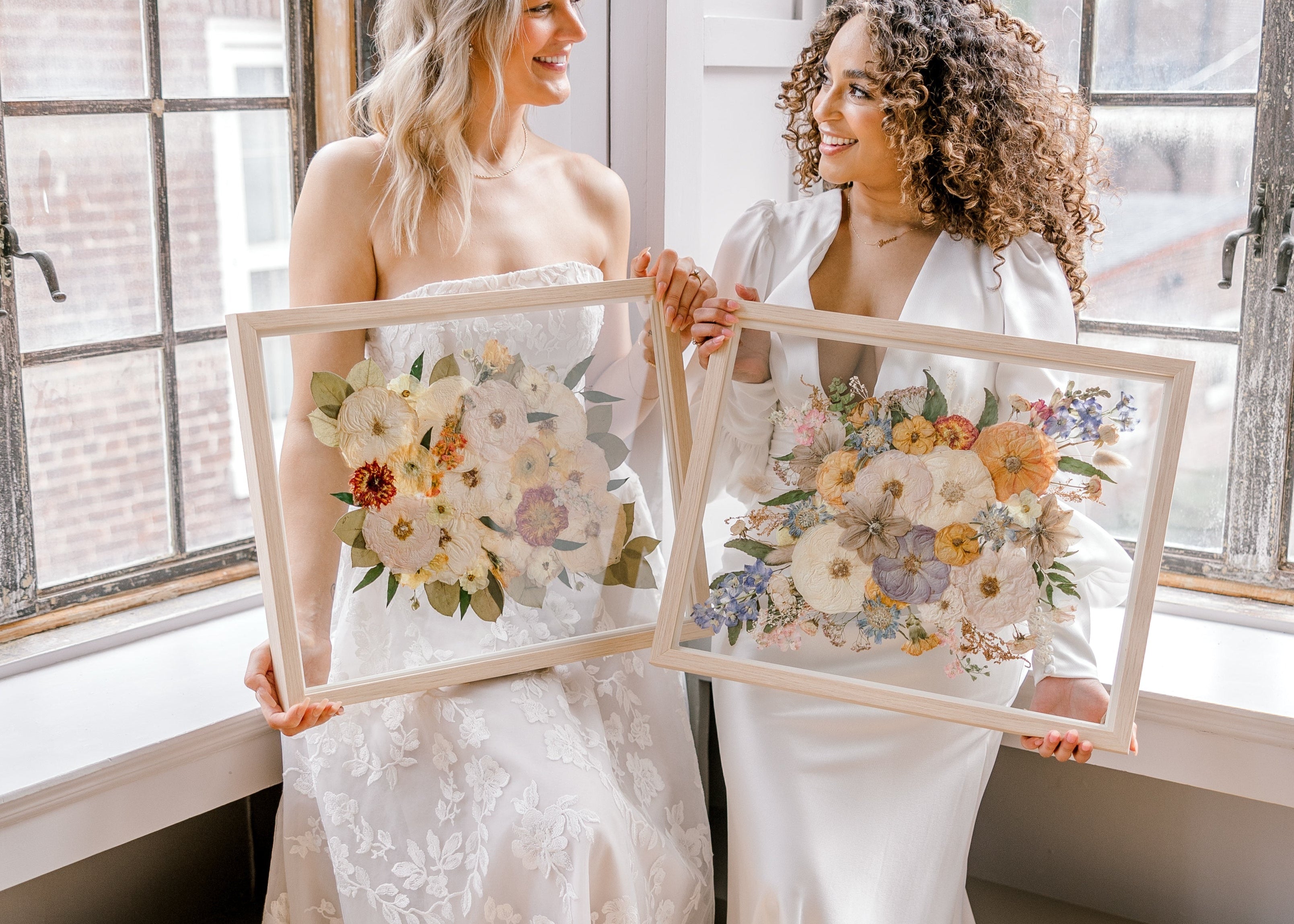 Two brides happily holding their pressed flower preservation pieces, smiling at each other and the lifetime of memories they will hold.
