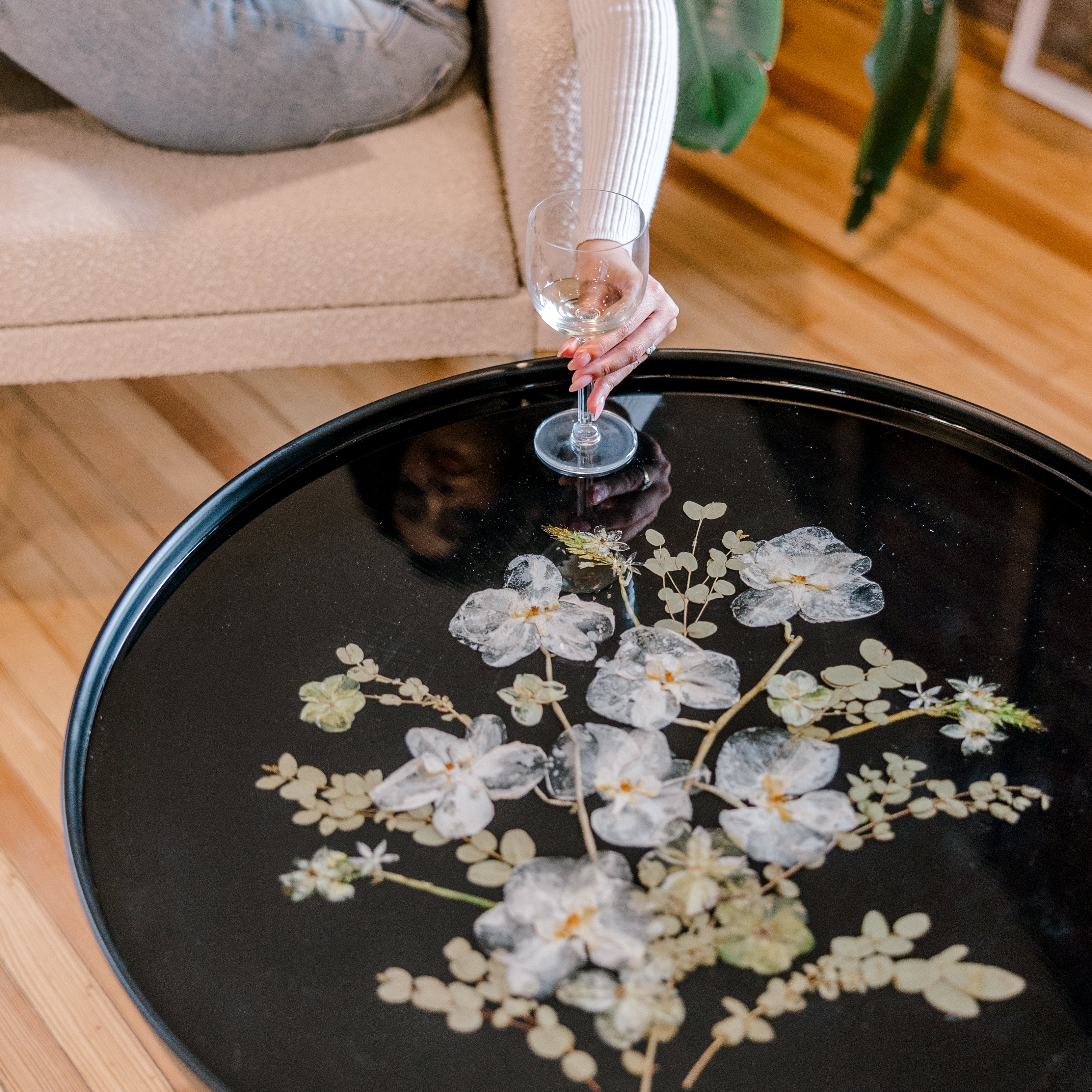 A person sitting in a white chair leaning over a resin table filled with pressed white florals grabbing a wine glass.