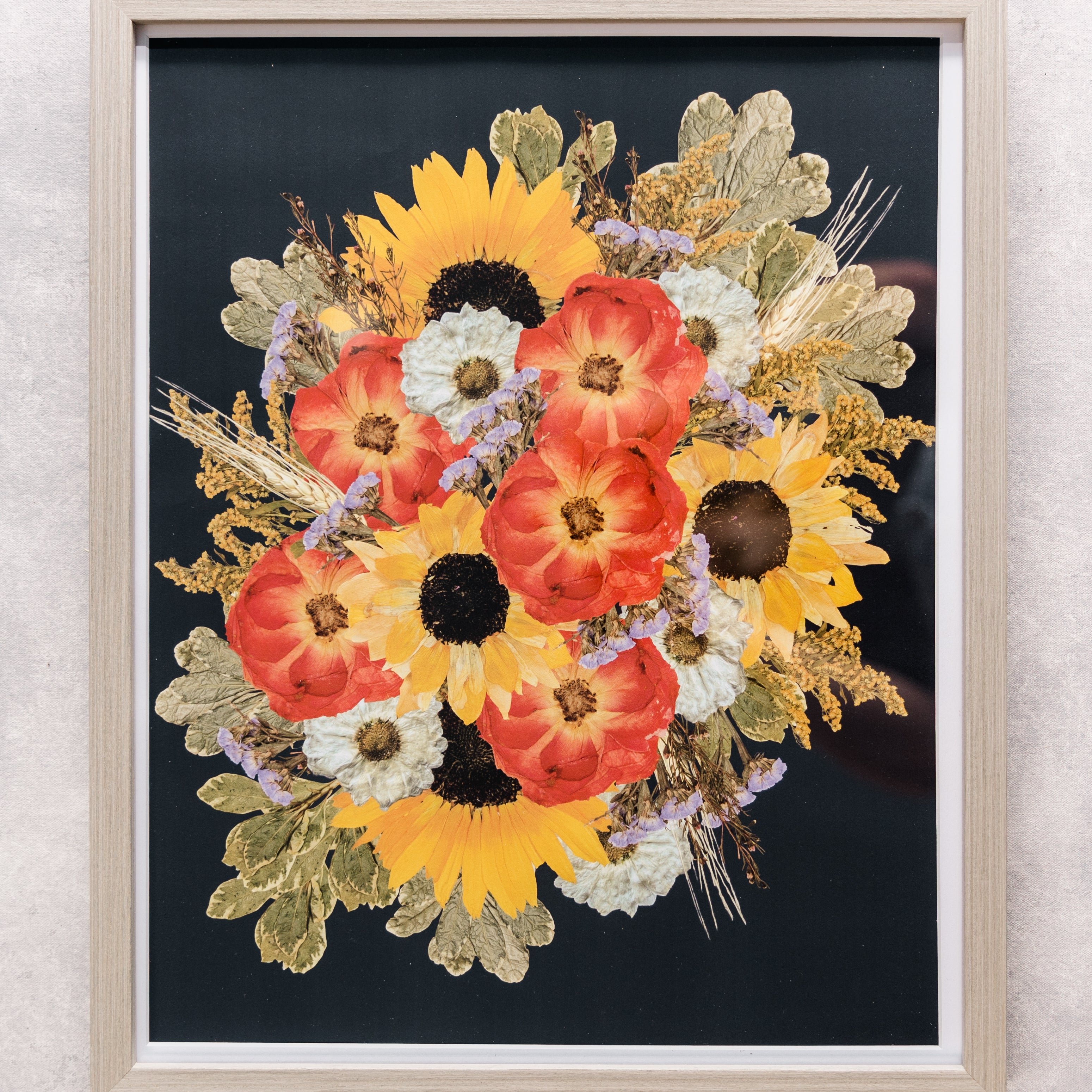 A large gray frame with sunflowers and pink or red pressed flowers against a navy blue background