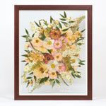 A large pressed floral bouquet encased in a brown frame with a date written in white script