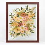 A large pressed floral bouquet encased in a brown frame with a date written in white script