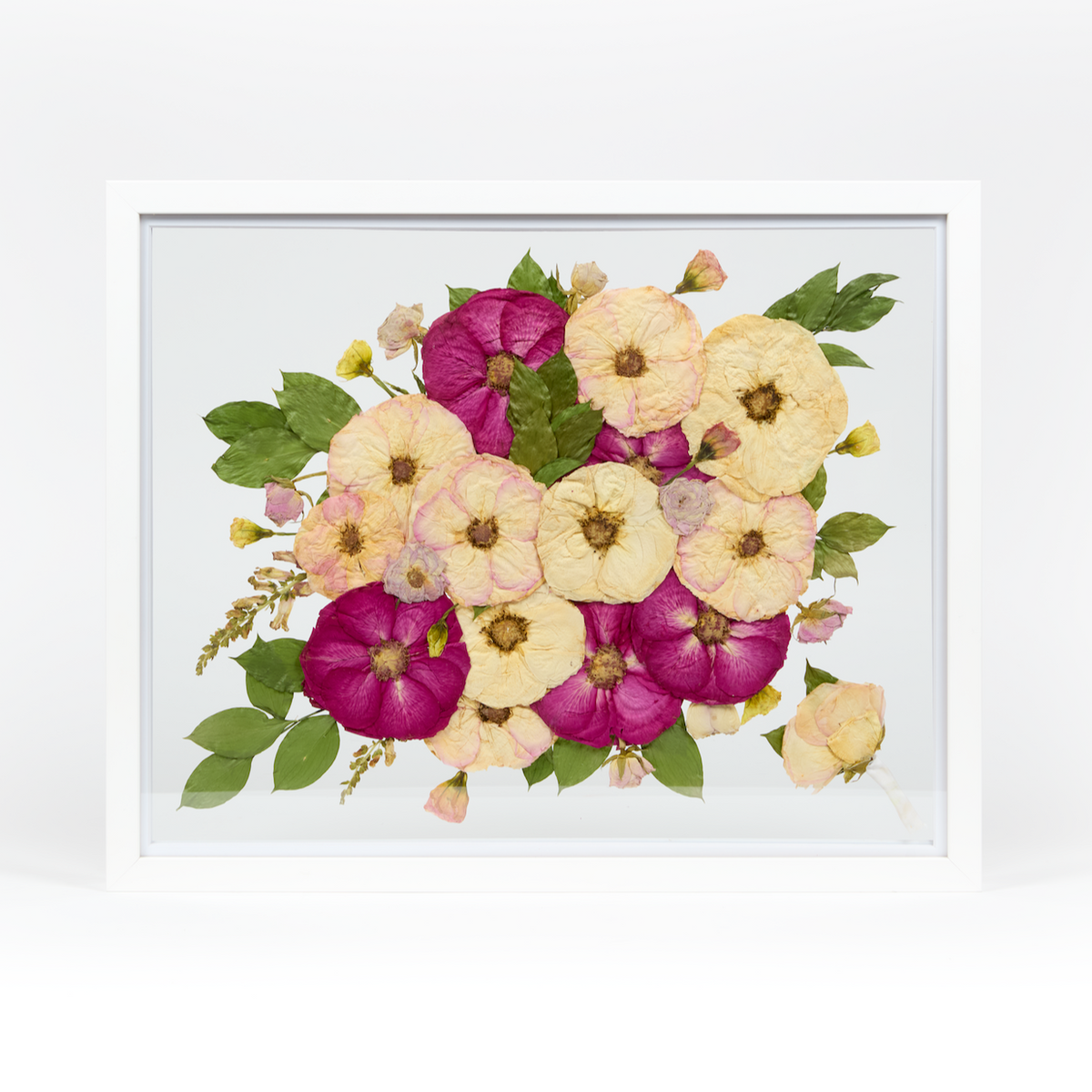 A pressed bridal bouquet in a white wood floating frame featuring pressed pink, red, and white florals with a preserved boutonniere in the corner.