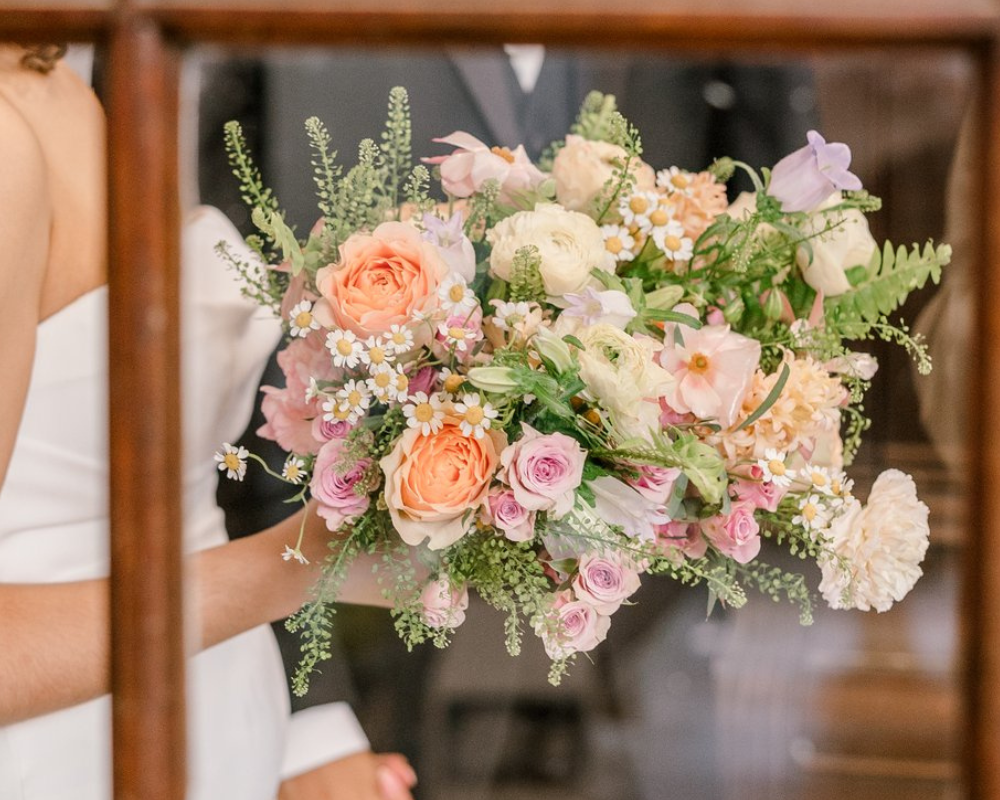 A bride is holding her wedding bouquet in one hand and her husband's hand in the other. Her bouquet includes white, pink, and orange flowers with greenery bursting from all sides.
