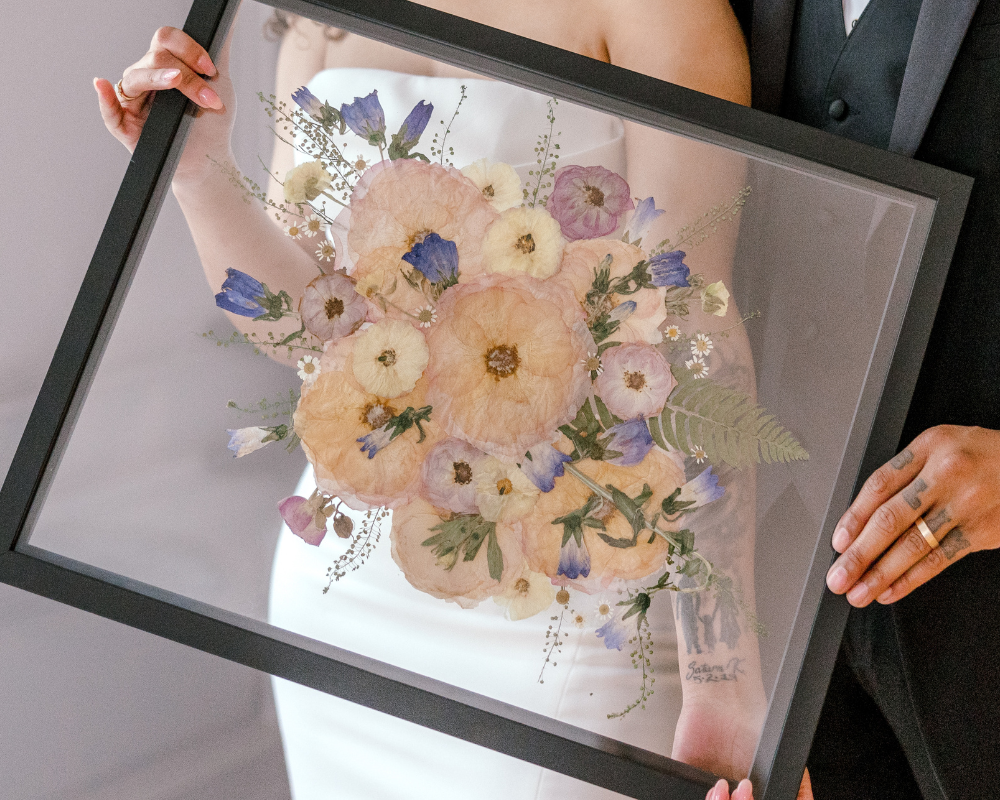 A newlywed couple is holding their pressed pink, blue, and white bouquet, preserved in a 16x20 black wood frame with a floating glass background.