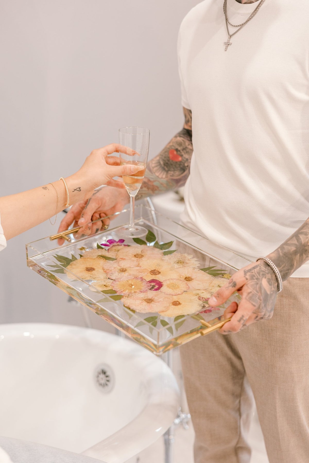 A man holds a tray of flowers as he hands his wife a glass of champagne in the bathtub.