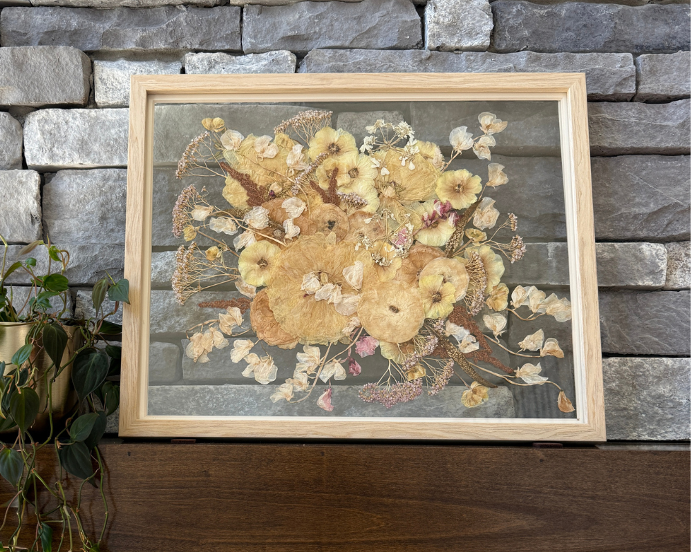 A pressed white bouquet in a 16x20 natural wood frame with glass backing is perched against a gray stone wall on a wooden mantel.