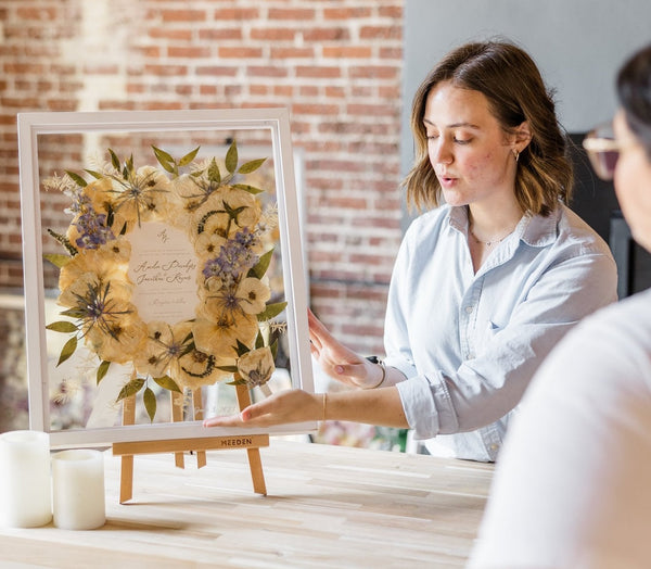 two women pointing and talking about a large white frame with pressed flowers
