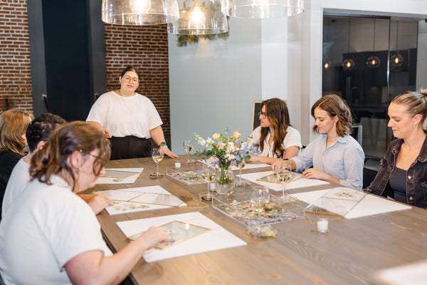 A women teaching a class o how to create a pressed flower frame at a large table with a bunch of people attending the workshop.