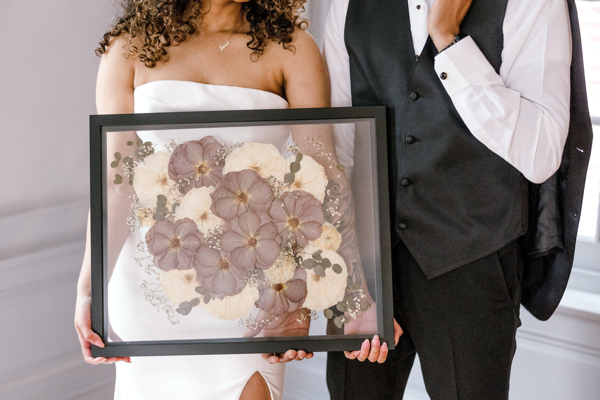 a bride holding a large black frame with pressed white and purple flowers next to a groom