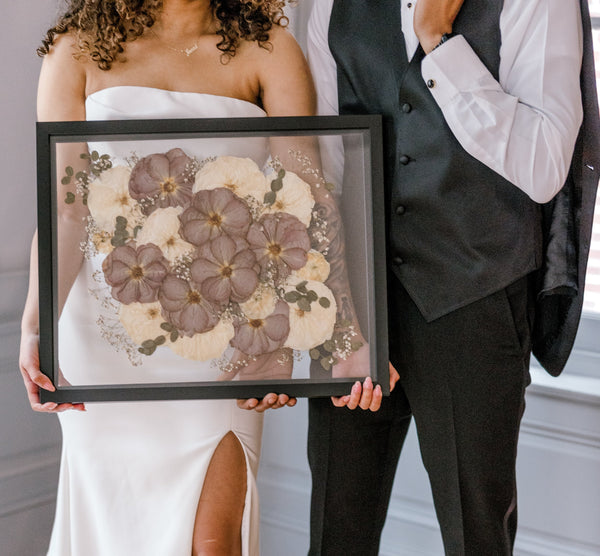 Bride and groom pose for a picture with a frame encased with pressed flowers from the special day in their home.