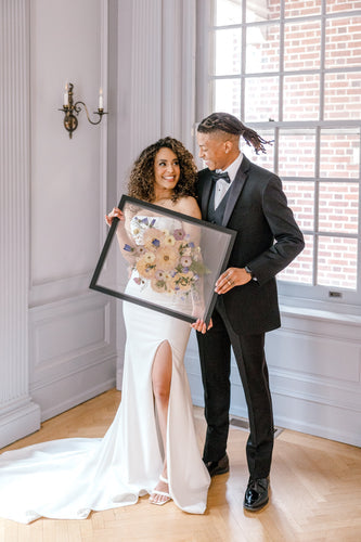 a bride and groom smiling at each other holding a large black frame with a colorful pressed bouquet encased