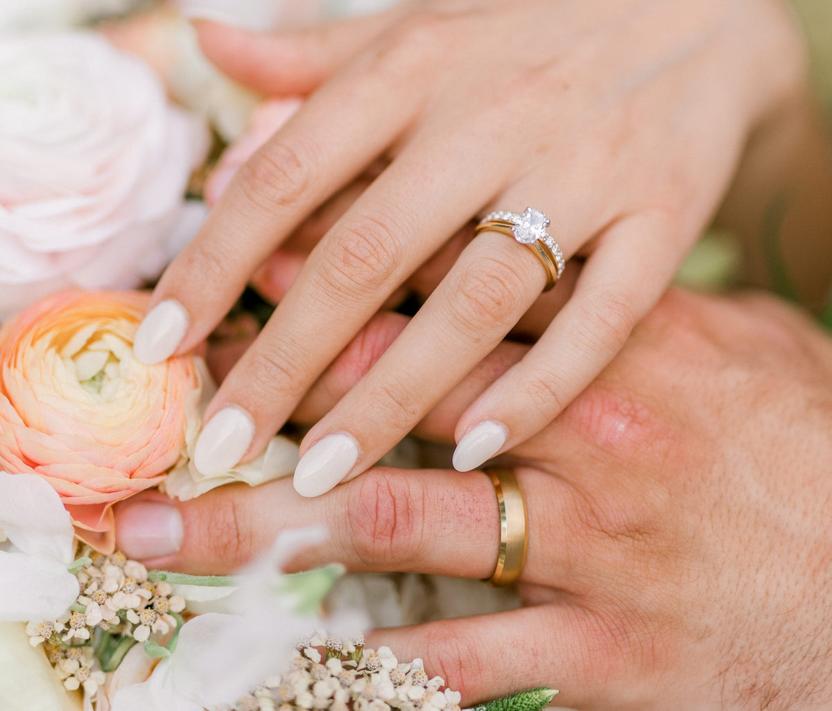 Close-up of a couple’s hands with wedding rings resting over a fresh bouquet of pink and orange flowers, capturing a joyful wedding moment.