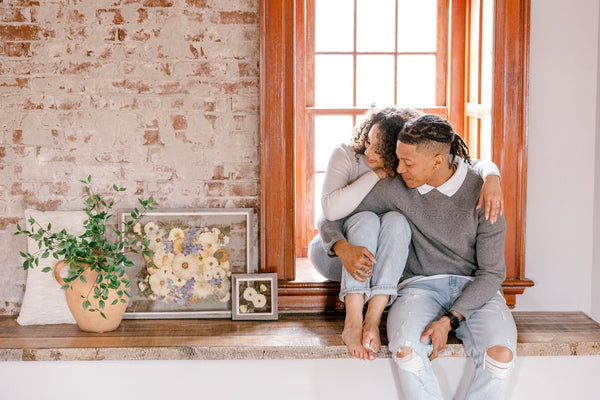 A couple sitting on a window sill admiring their pressed bouquet frames lovingly. 