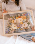 Beautiful pressed florals displayed in a floating glass frame held by a bride admiring her bouquet preservation.