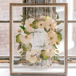 A beautiful bouquet preservation featuring white pressed roses and a preserved wedding invitation surrounded by pressed greenery in a barn wood frame.