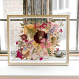 Brightly colored pressed florals are featured in this preserved bridal bouquet. The pressed flower frame is displayed in a gold wood frame against rustic windows.