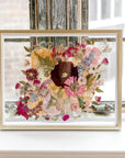 Brightly colored pressed florals are featured in this preserved bridal bouquet. The pressed flower frame is displayed in a gold wood frame against rustic windows.