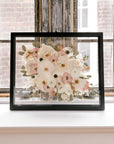 A pressed floral bouquet frame with white, pink, and green pressed flowers. Showcased in a black wood floating frame, this bouquet preservation shines beautifully in its forever home.