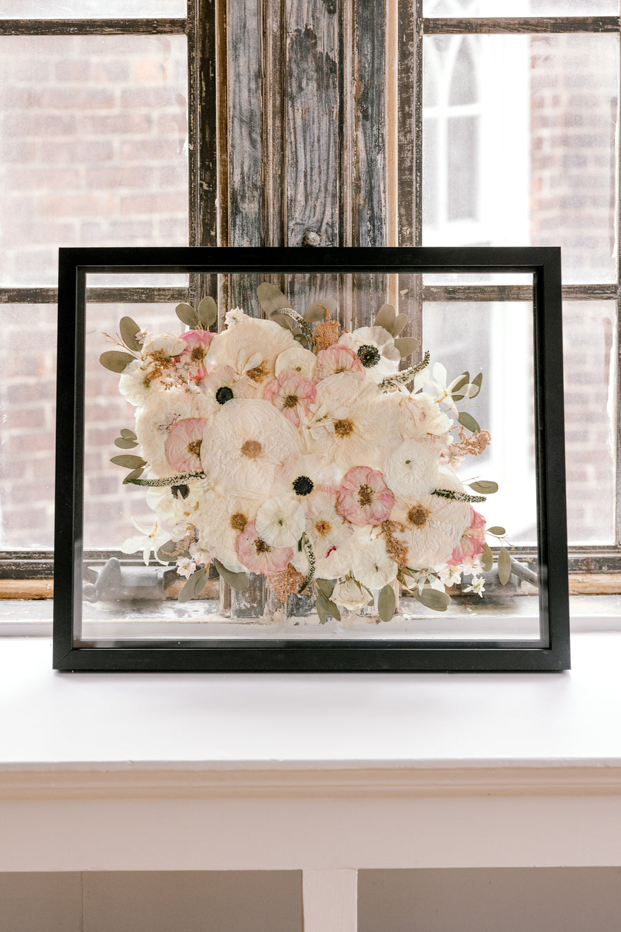 A pressed floral bouquet frame with white, pink, and green pressed flowers. Showcased in a black wood floating frame, this bouquet preservation shines beautifully in its forever home.