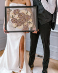 A happy couple stands holding their wedding bouquet preservation featuring pressed white roses and pressed purple roses with accents of pressed baby's breath and pressed eucalyptus. 