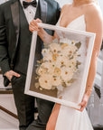 A happy couple admiring their bouquet preservation piece featuring white pressed flowers and pressed greenery accents in a white wood floating frame. 