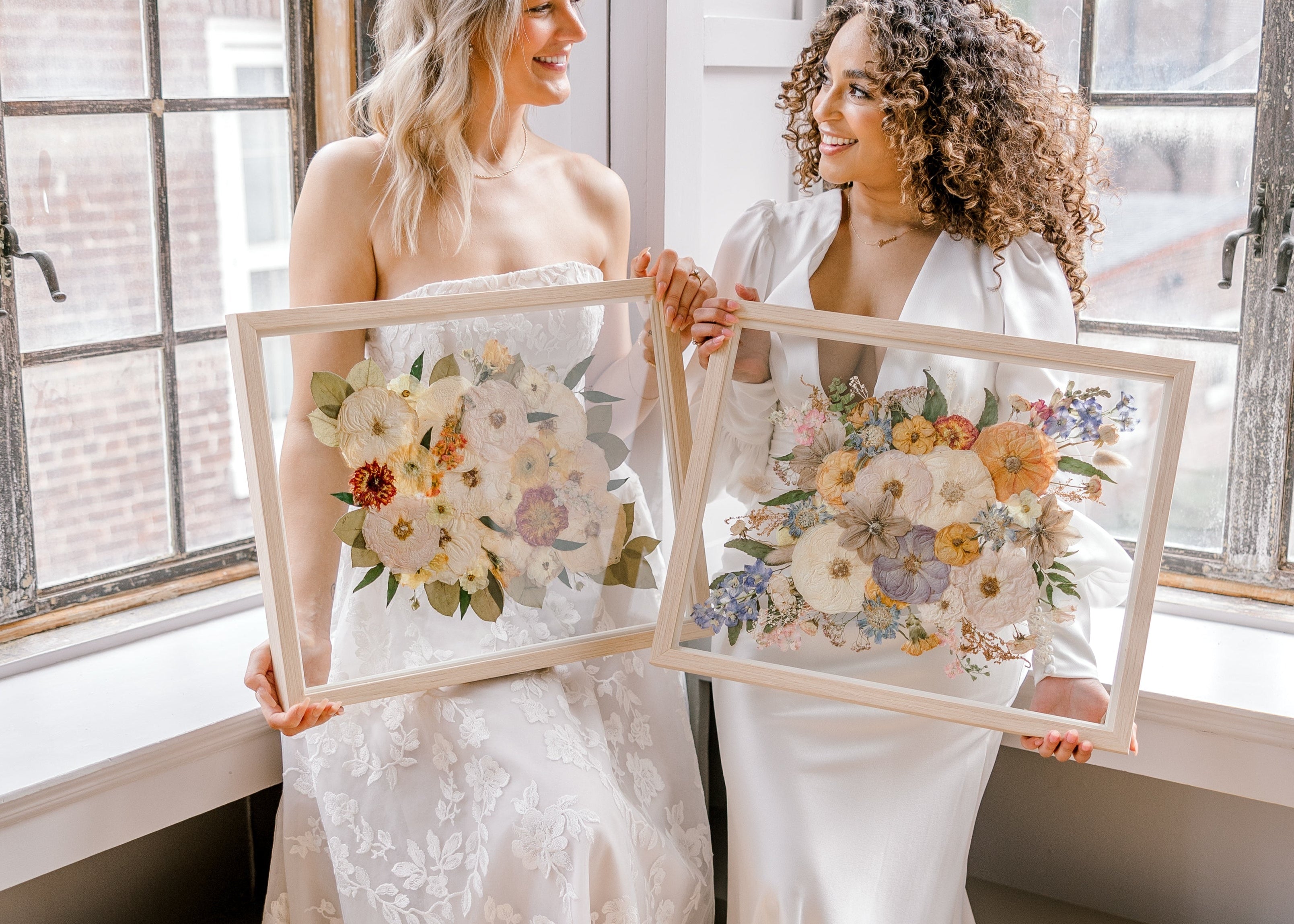 Two brides happily holding their pressed flower preservation pieces, smiling at each other and the lifetime of memories they will hold. 