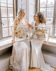 Two brides happily holding their pressed flower preservation pieces, smiling at each other and the lifetime of memories they will hold. 