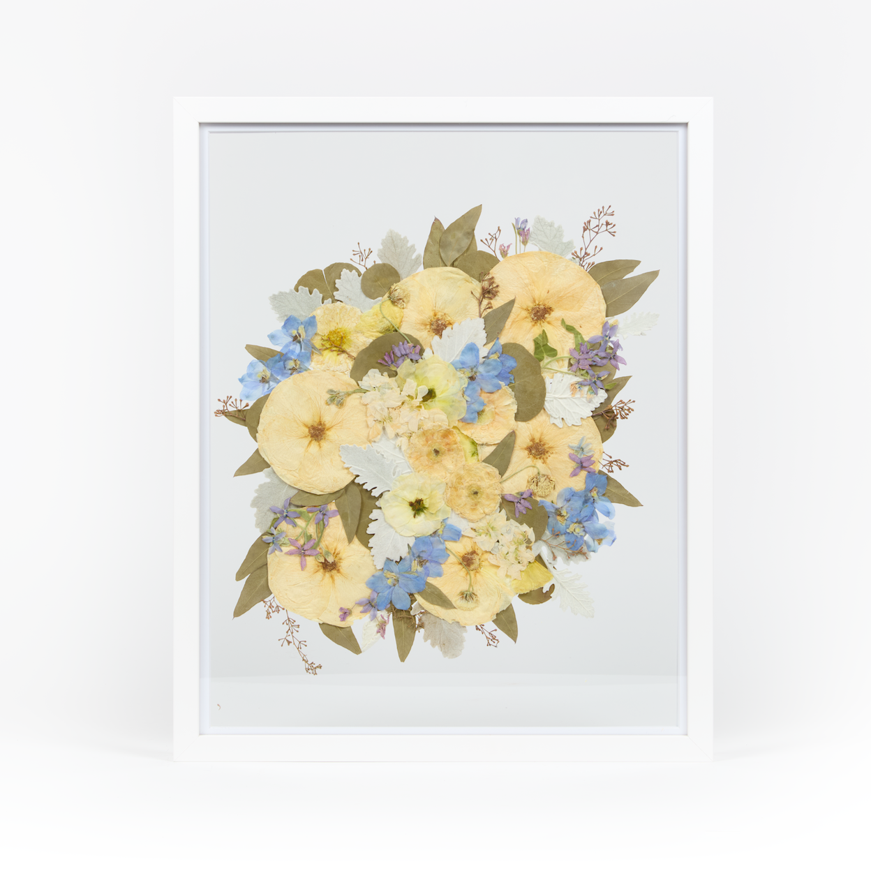 A pressed bridal bouquet in a white wood floating frame featuring pressed blue and white florals. 