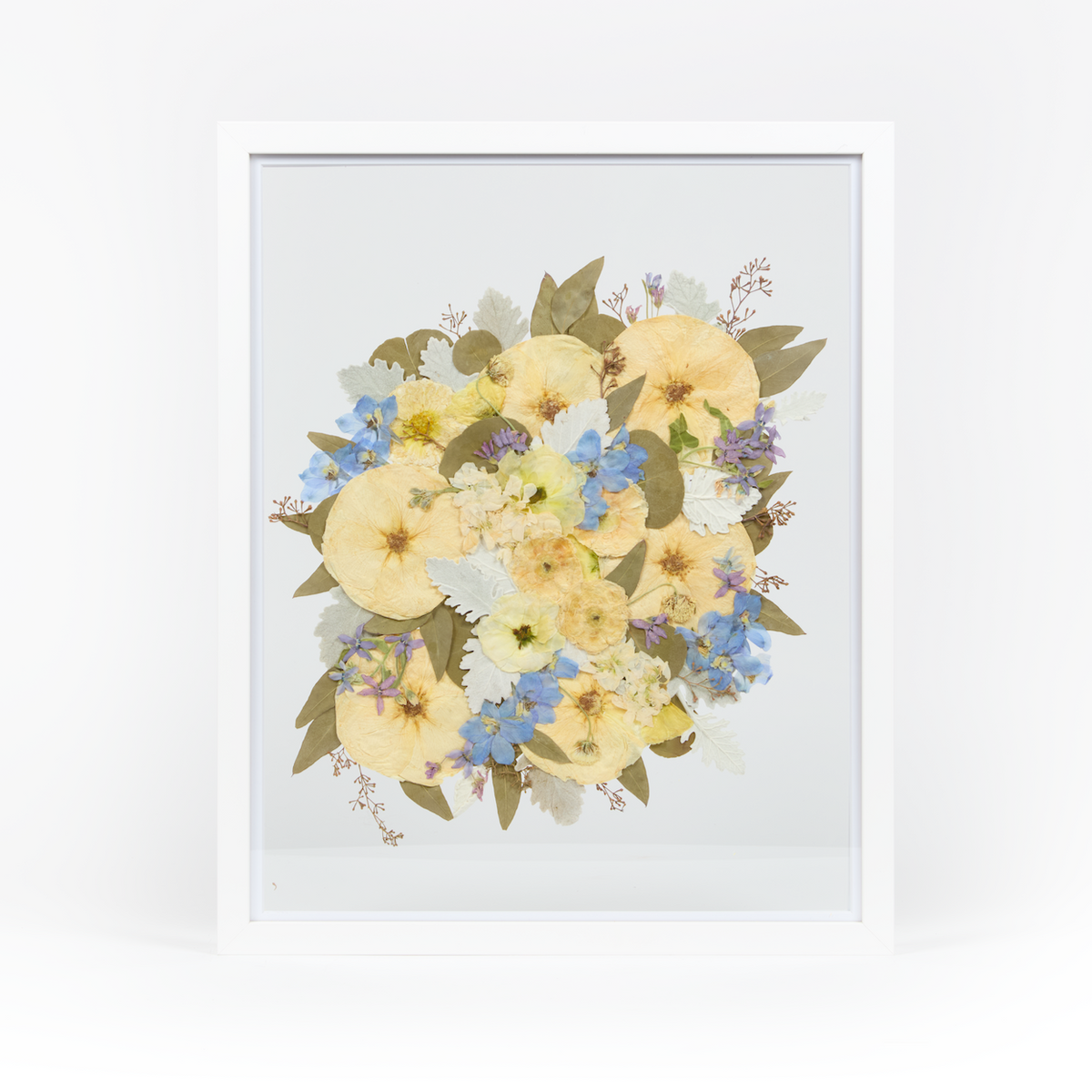 A pressed bridal bouquet in a white wood floating frame featuring pressed blue and white florals. 