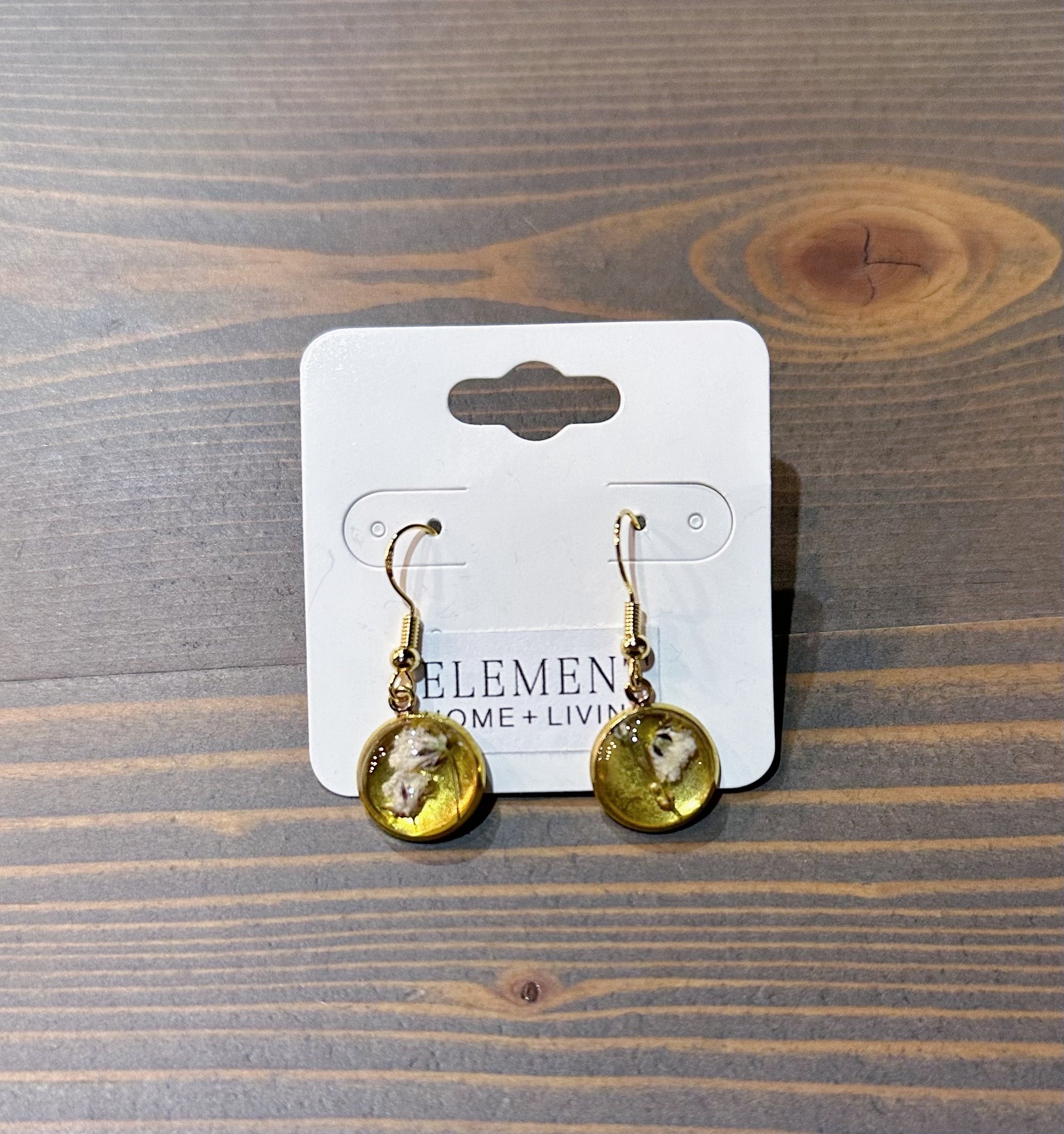 A pair of gold dangle earrings feature real pressed white flowers in a circular gold bezel setting. They are set against a wooden background in a white earring holder.