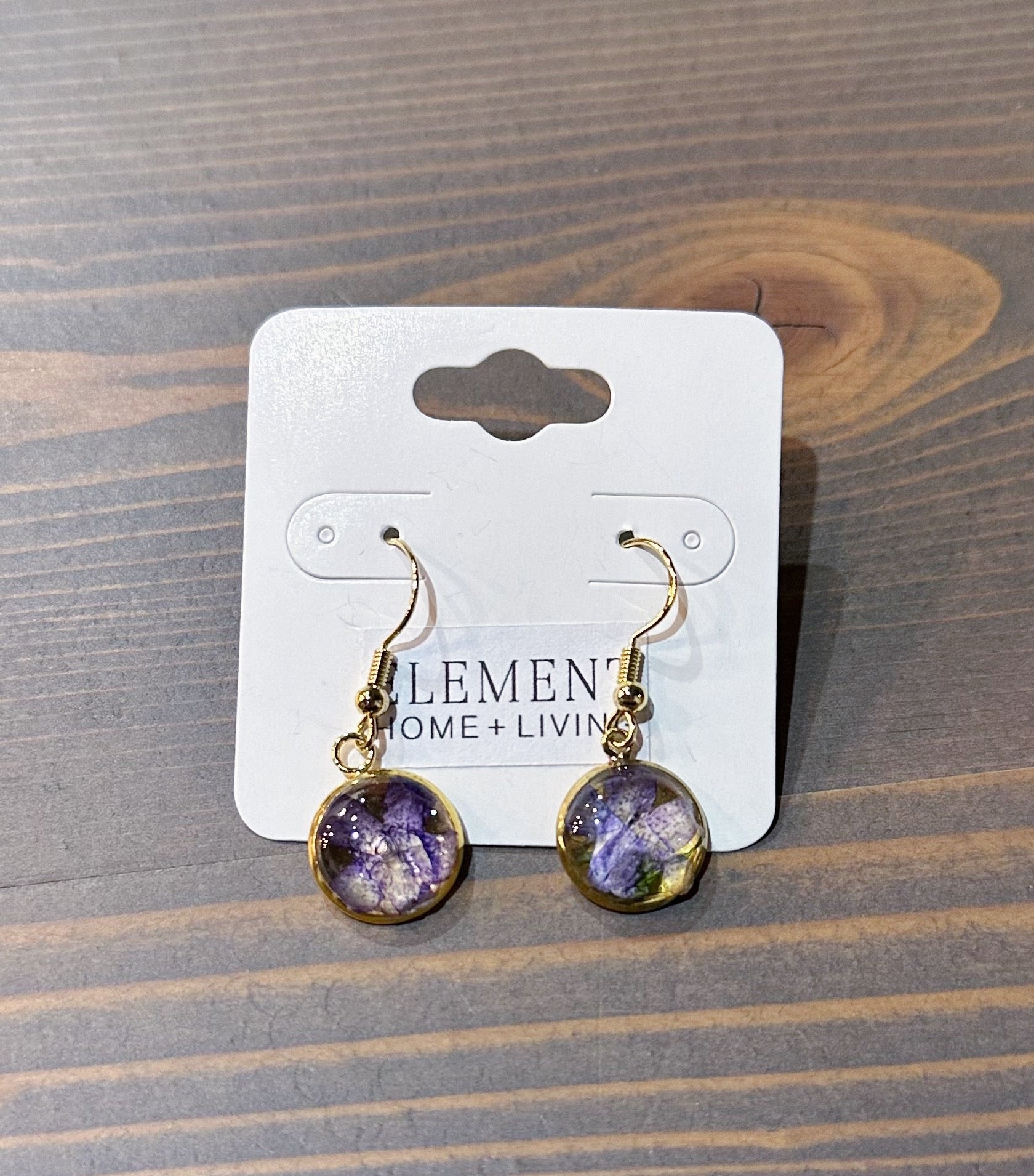 A pair of circular gold dangle earrings feature real pressed purple flowers. They are placed on top of a wooden surface. 