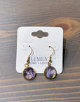 A pair of circular gold dangle earrings feature real pressed purple flowers. They are placed on top of a wooden surface. 