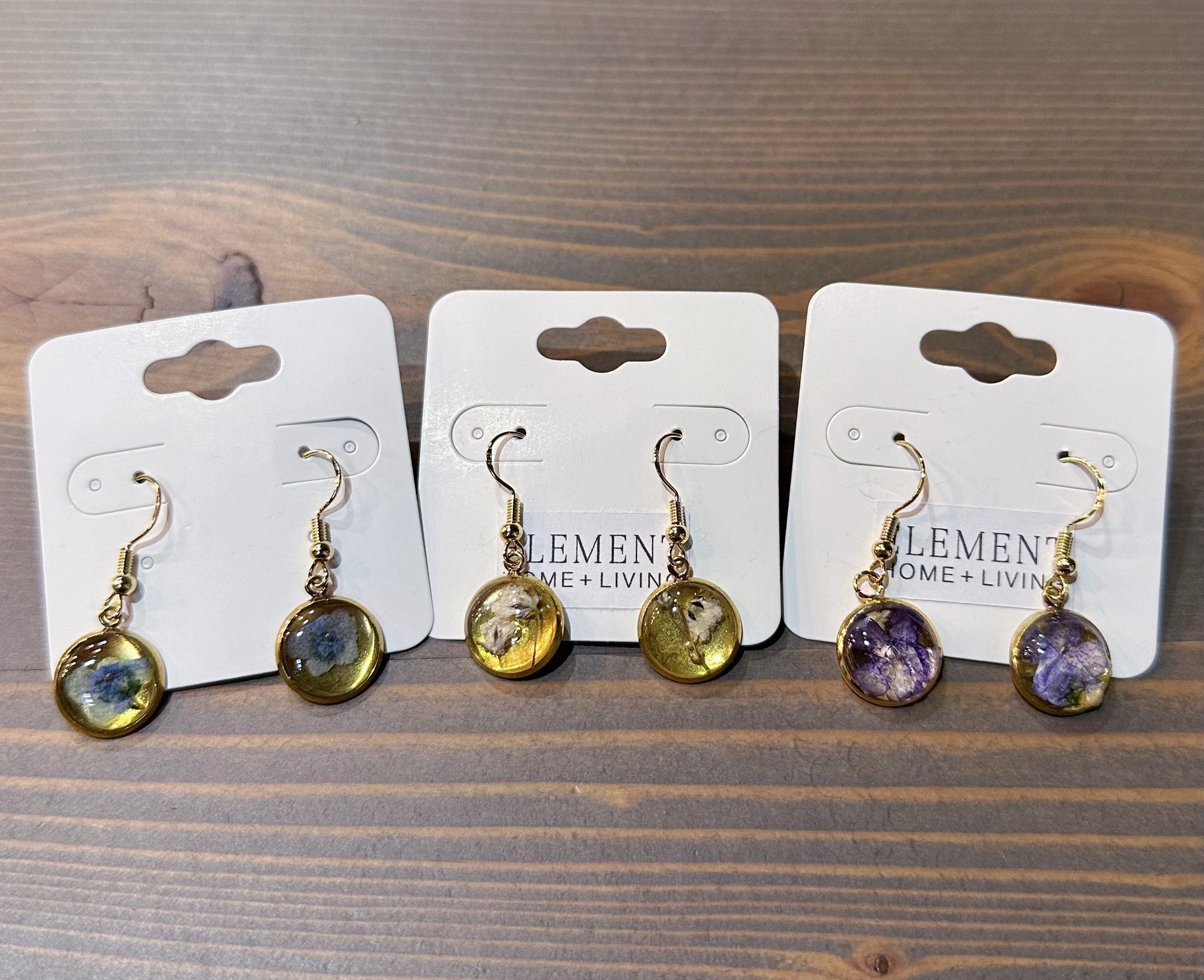 Three pairs of gold dangle resin earrings are placed in a line across a wooden surface. Each pair features a different color pressed flower: blue, white, and purple. 