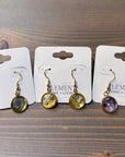 Three pairs of gold dangle resin earrings are placed in a line across a wooden surface. Each pair features a different color pressed flower: blue, white, and purple. 