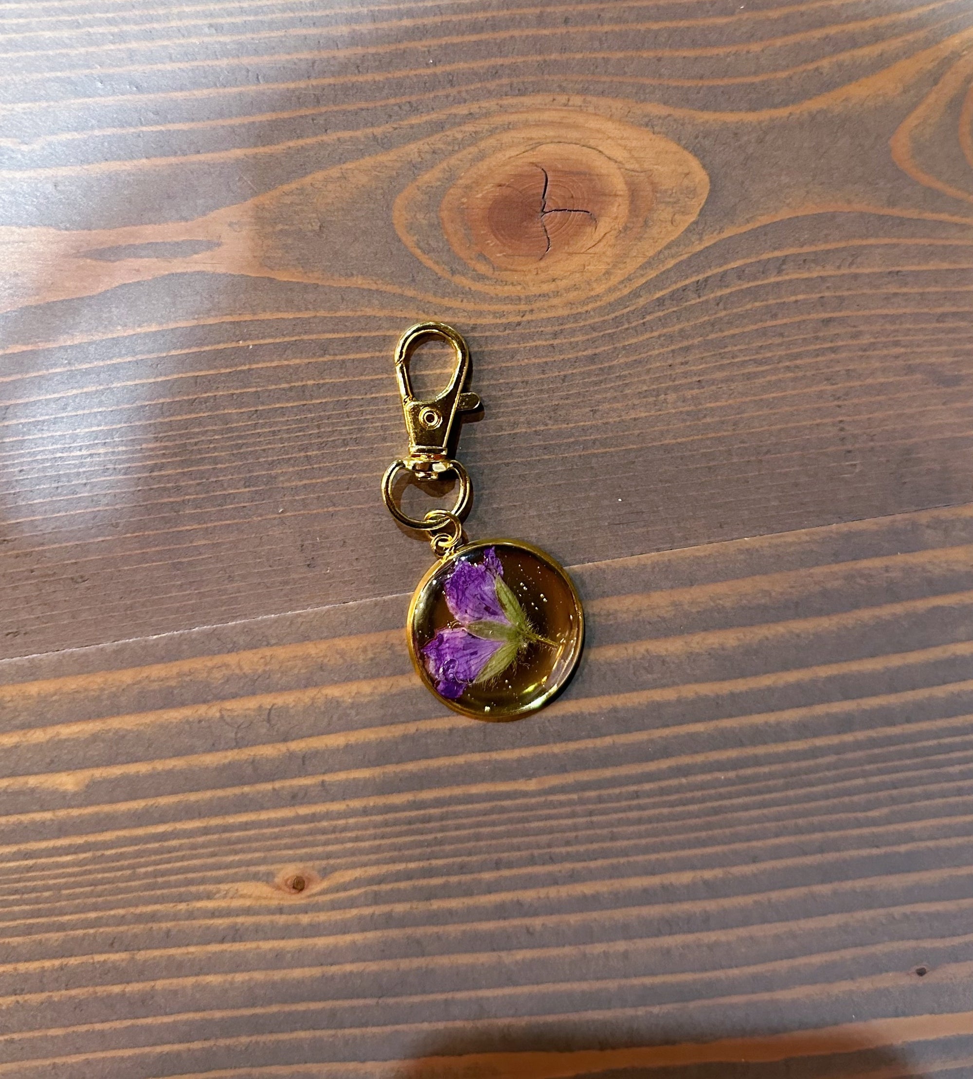 A gold pressed flower keychain includes one small purple flower. It is placed against a wooden background. 