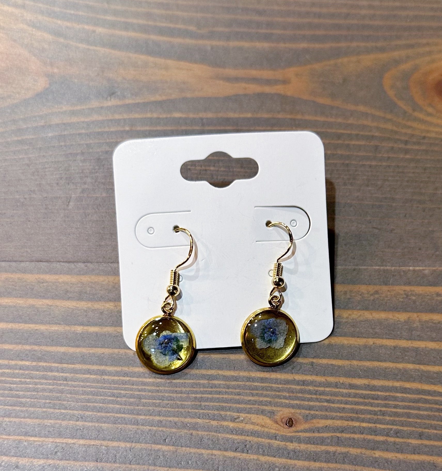 A pair of gold dangle earrings feature real pressed blue flowers within a circular gold bezel setting. They are placed on top of a wooden surface. 