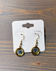 A pair of gold dangle earrings feature real pressed blue flowers within a circular gold bezel setting. They are placed on top of a wooden surface. 