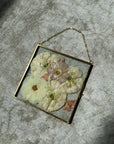 a small square gold hanging frame with pressed flowers against a white rug.