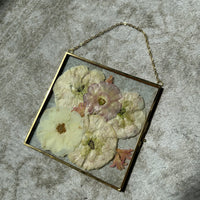 a small square gold hanging frame with pressed flowers against a white rug.