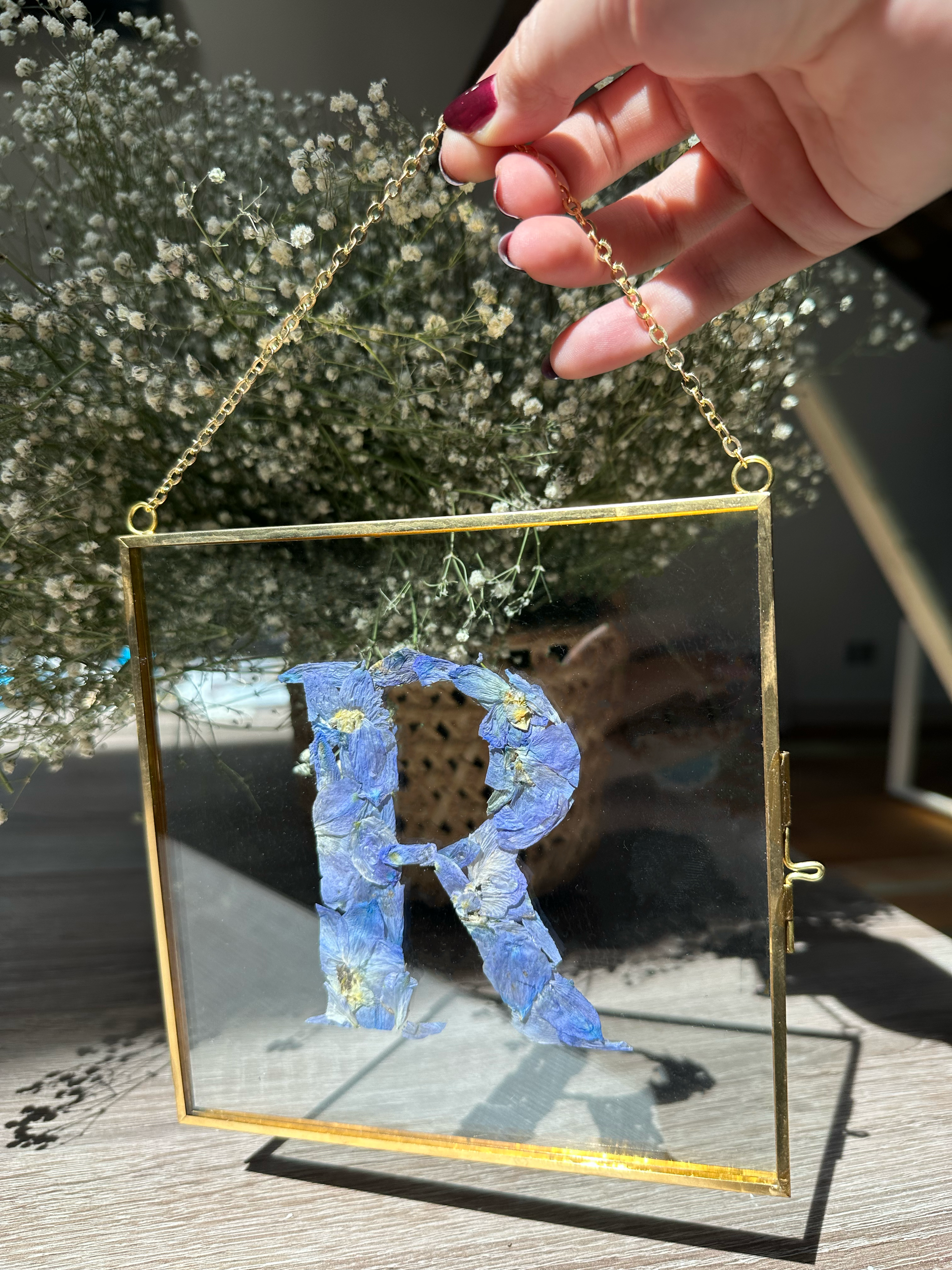 Gold hanging frame with pressed flower petals designed into the letter r held by a person.