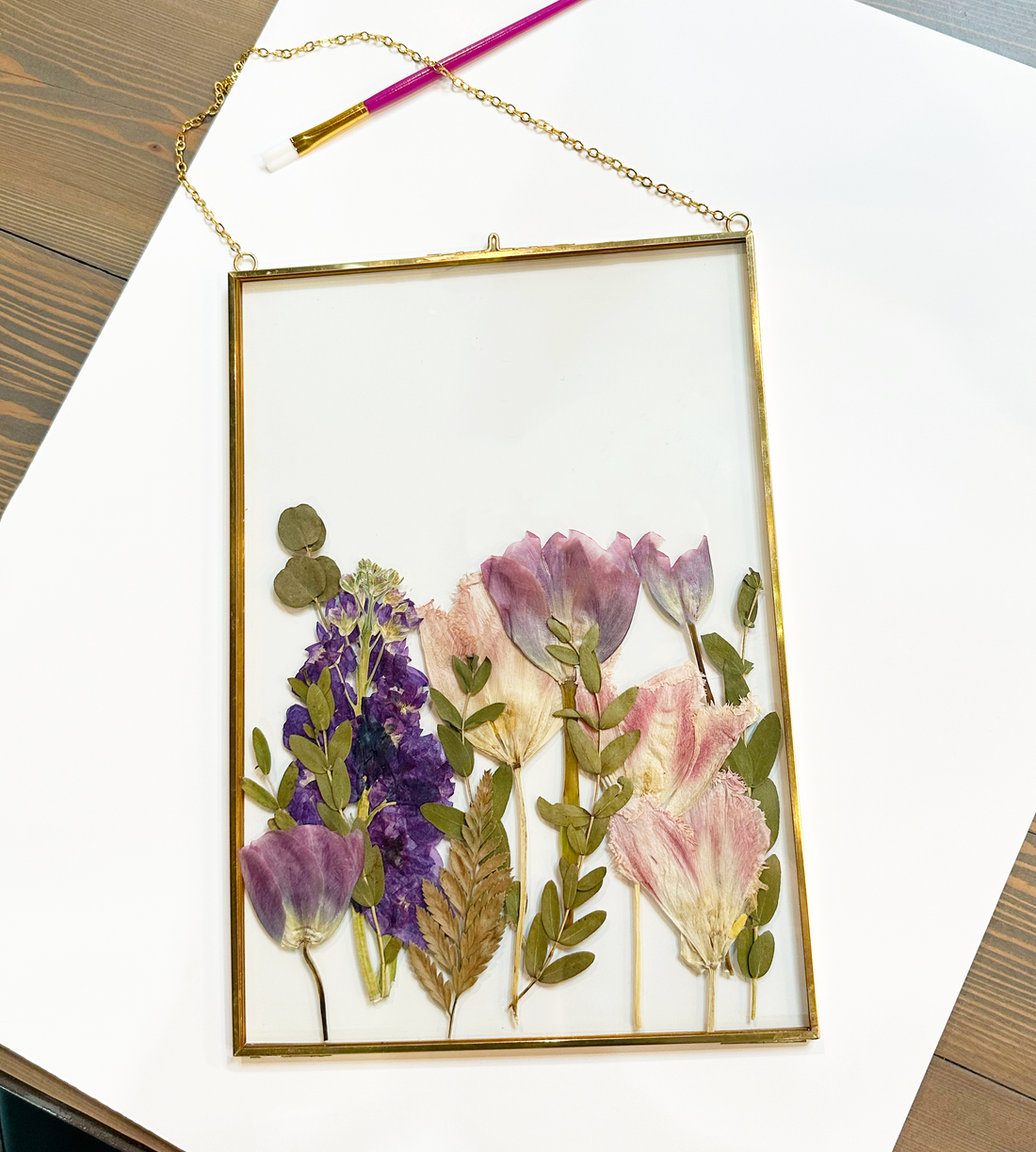 Purple pressed flowers inside a gold hanging frame on top of a white mat against a wooden table.