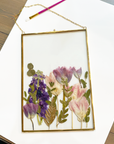 Purple pressed flowers inside a gold hanging frame on top of a white mat against a wooden table.