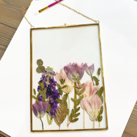 Purple pressed flowers inside a gold hanging frame on top of a white mat against a wooden table.