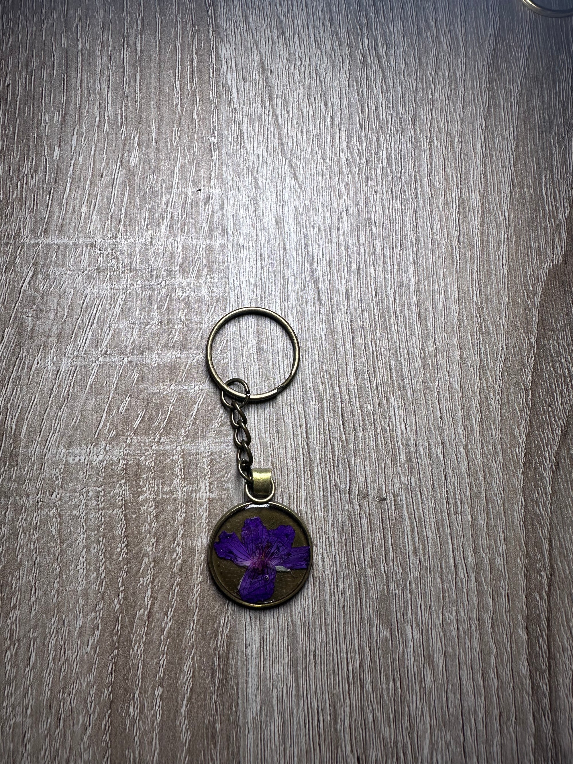 A bronze pressed flower keychain with one small purple flower is placed against a wooden background. 