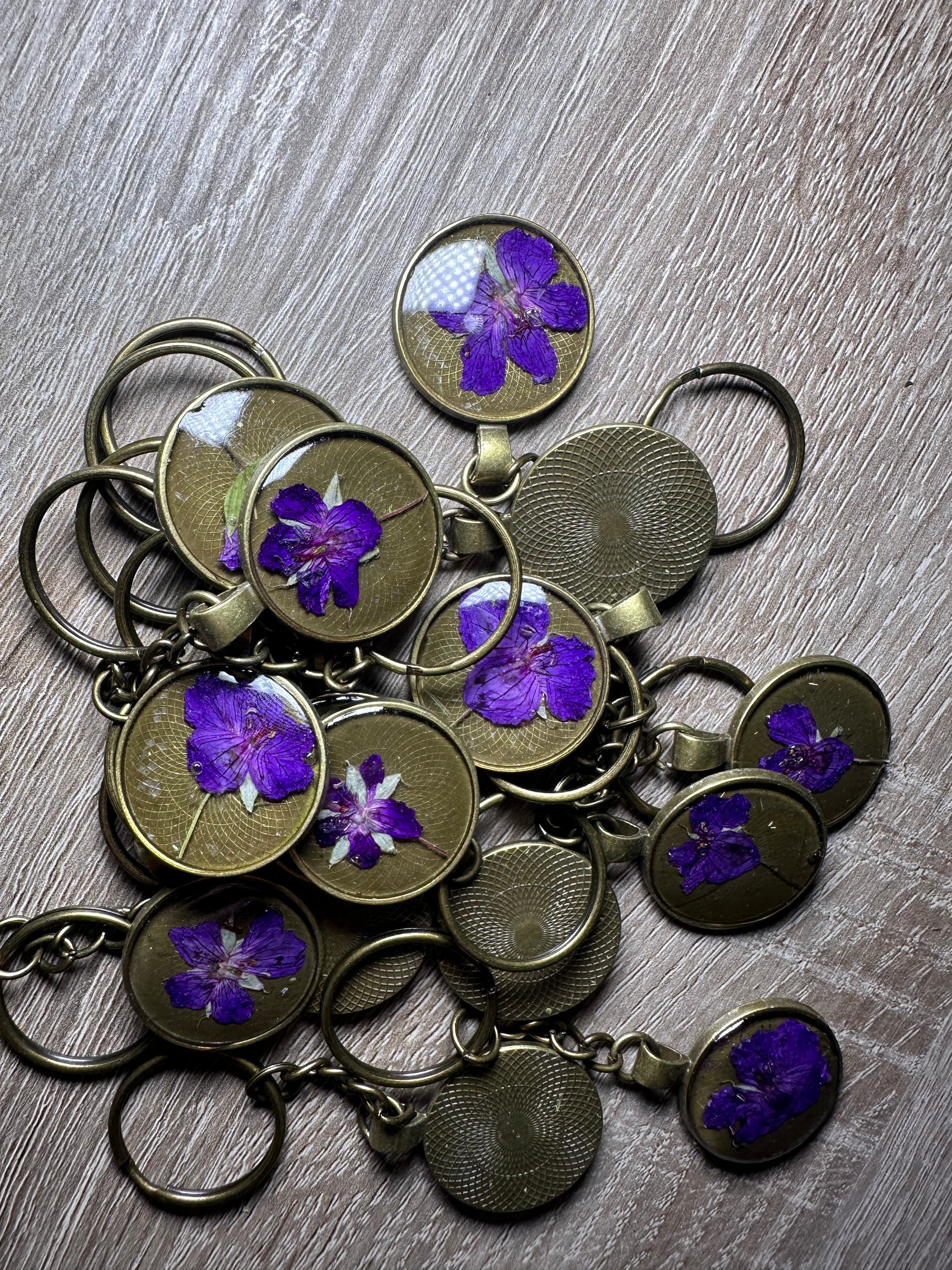 A small pile of bronze pressed flower keychains, with one purple flower in each, is placed on top of a wooden surface. 