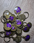 A small pile of bronze pressed flower keychains, with one purple flower in each, is placed on top of a wooden surface. 