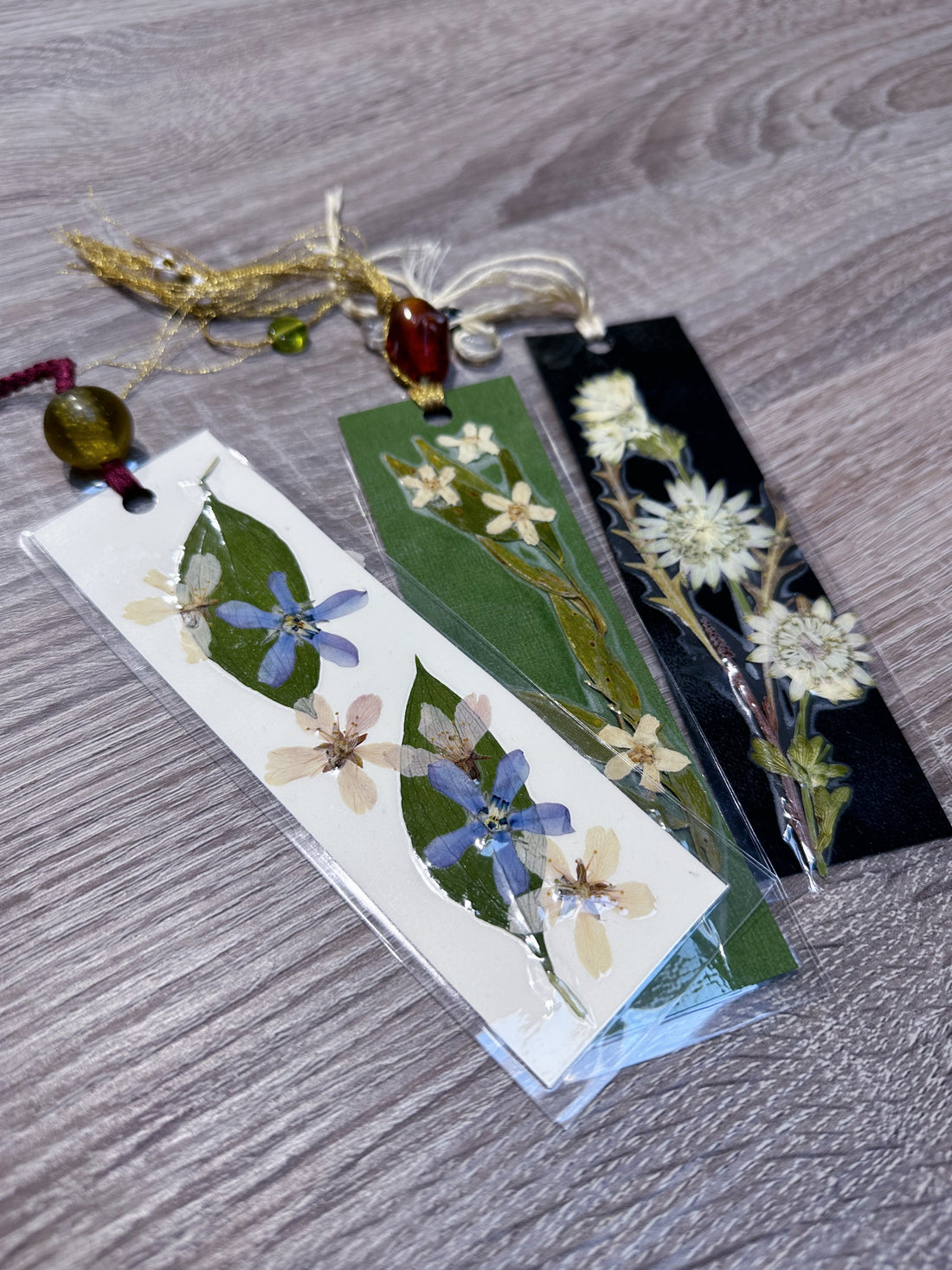 Three bookmarks on a wooden table with pressed flowers and attached by a string and beads