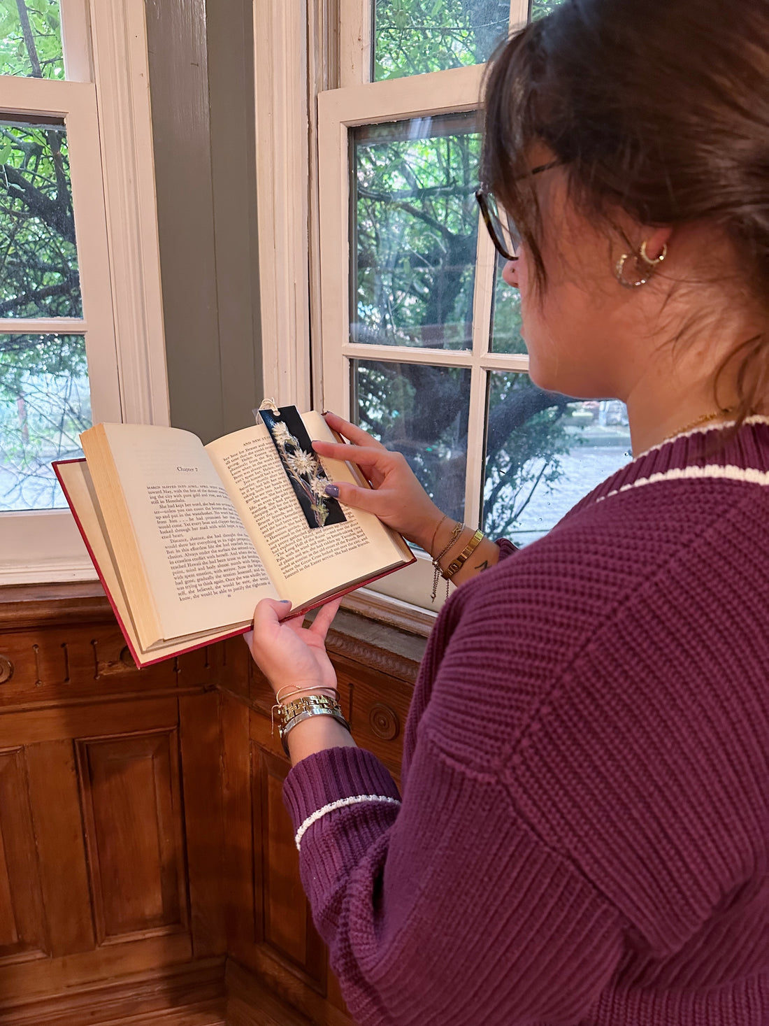 A woman reading a book holding a pressed flower bookmark to hold open the page