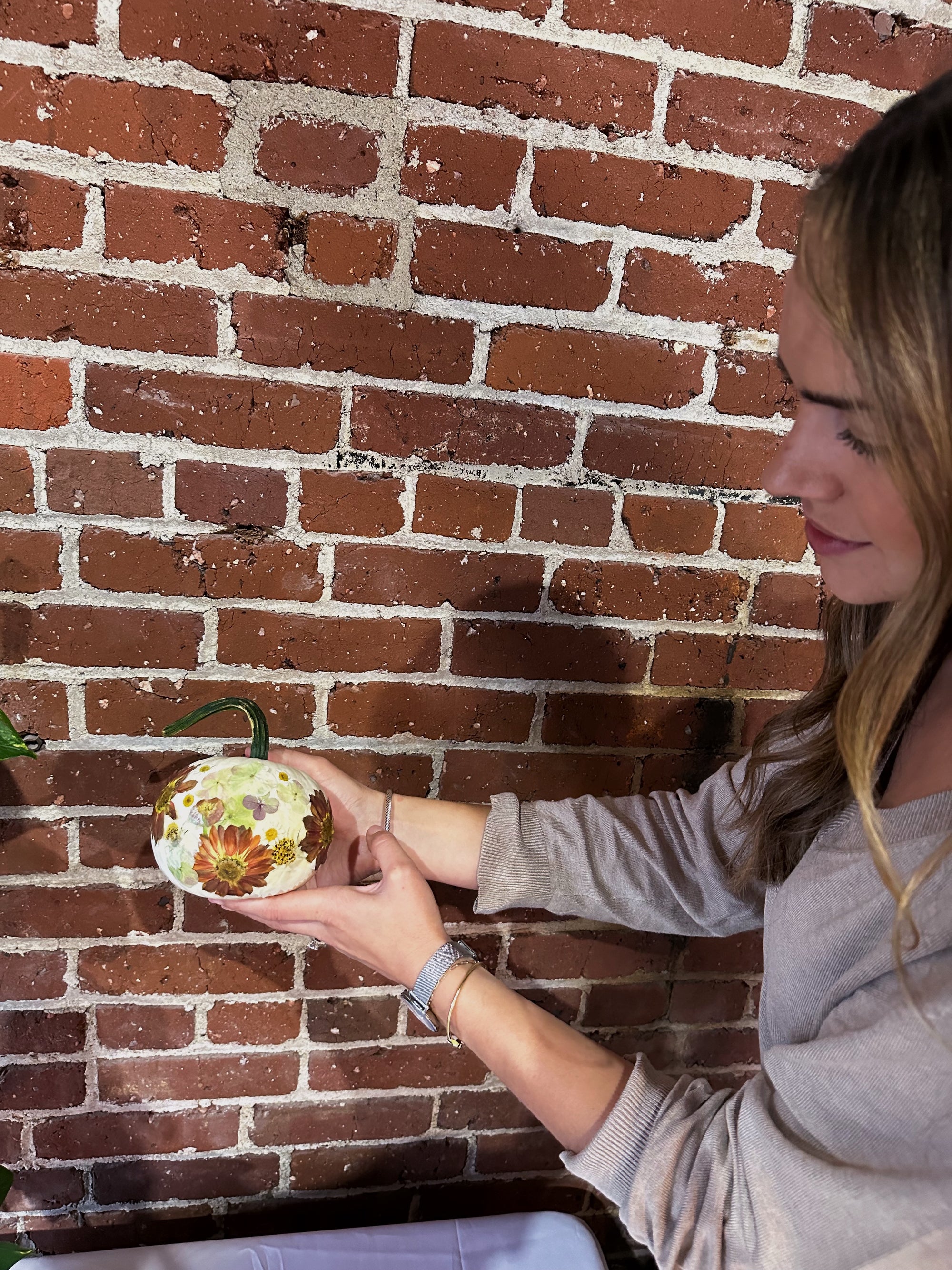 a woman holding a small white pumpkin designed with pressed florals in front of a brick wall